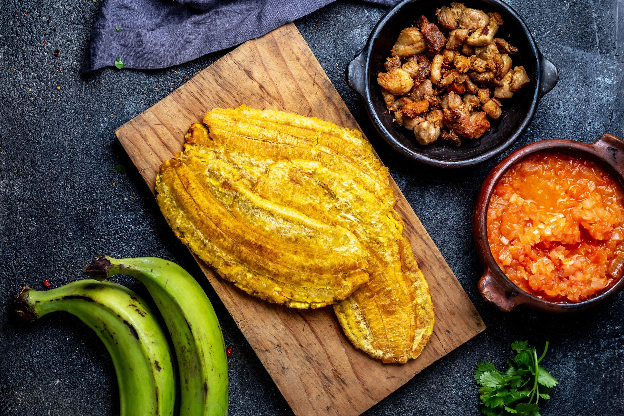 COLOMBIAN CARIBBEAN CENTRAL AMERICAN FOOD. Patacon or toston, fried and flattened whole green plantain banana on white plate with tomato sauce and chicharron Black background, top view | GreySuits Advisors | GreySuits Advisors