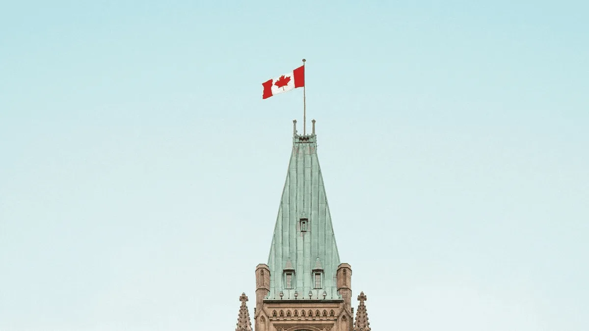 Flag atop a tall Gothic spire: Canada flag and a red flag fly over a historic stone tower against a pale blue sky.