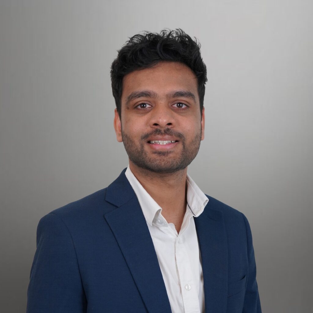 Varshil Headshot | GreySuits Advisors | GreySuits Advisors Portrait of a man in a navy blazer and white shirt, smiling at the camera against a gray background.