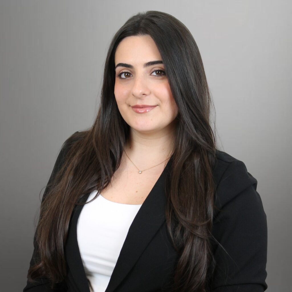 Zarmig Headshot | GreySuits Advisors | GreySuits Advisors Professional headshot of a woman with long dark hair, wearing a black blazer and white top, against a neutral gray background.