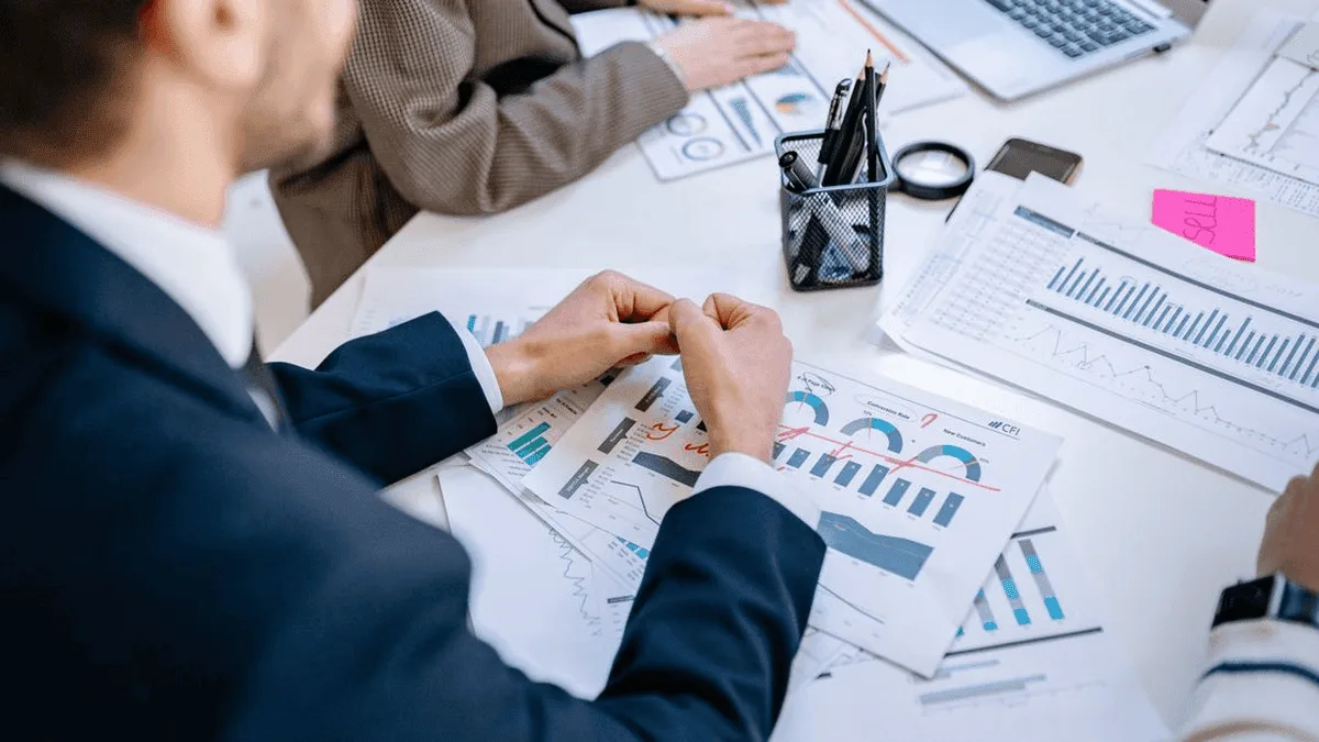Team members examine printed charts and graphs on a white table during a meeting.