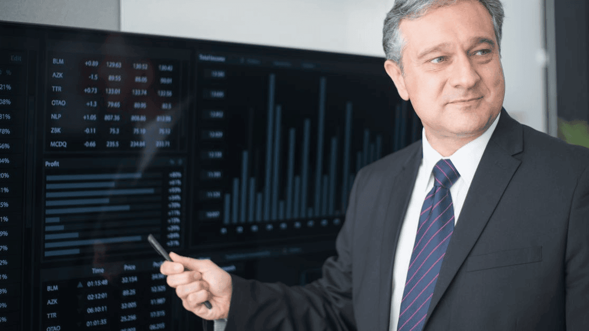 GreySuits-Feature-pexels-photo-8353807-104 | GreySuits Advisors | GreySuits Advisors Professional man in a dark suit points at financial charts on large monitor walls displaying stock data and graphs behind him
