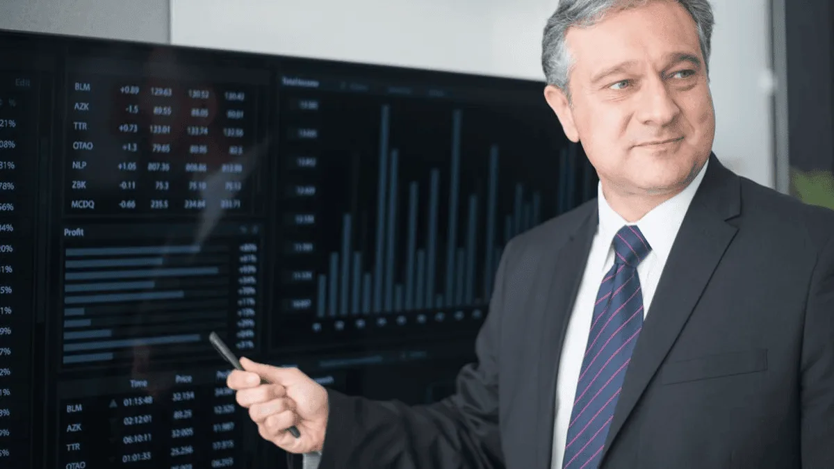Professional man in a dark suit points at financial charts on large monitor walls displaying stock data and graphs behind him