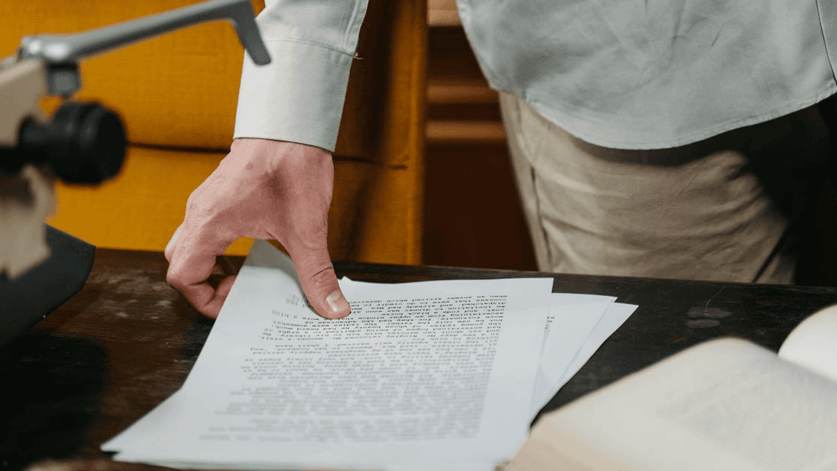 GreySuits-Feature-pexels-photo-9572697-652 | GreySuits Advisors | GreySuits Advisors Close-up of a hand turning the top page of a document on a dark desk, with an open book nearby.