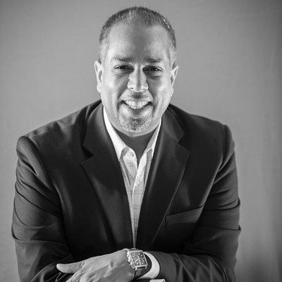 Professional man in a dark suit smiles at the camera, arms crossed, wearing a wristwatch (black-and-white portrait).