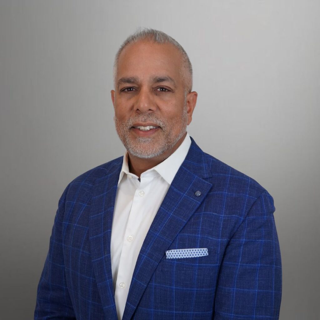 Professional head-and-shoulders portrait of a smiling man with short gray hair and a trimmed beard, wearing a blue plaid blazer and white shirt against a gray backdrop.